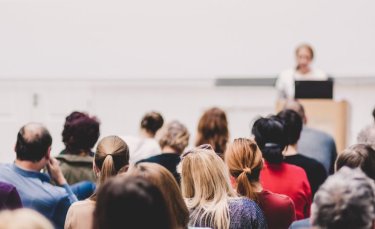 au premier plan, des hommes et des femmes sont assis dans une salle de conférence. Ils écoutent une femme debout derrière un pupitre